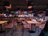 A modern cafeteria with neatly organized rows of tables and chairs, featuring a prominent illuminated sign that says 'LOVE AT FIRST BITE.' The seating area consists of light wooden tables and chairs with a mesh design. The ambiance is enhanced by pendant lights hanging from the ceiling, and there are large screens displaying images in the background.