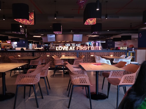 A modern cafeteria with neatly organized rows of tables and chairs, featuring a prominent illuminated sign that says 'LOVE AT FIRST BITE.' The seating area consists of light wooden tables and chairs with a mesh design. The ambiance is enhanced by pendant lights hanging from the ceiling, and there are large screens displaying images in the background.