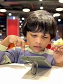 A young child attentively gazes at a tablet placed on a table, with a background that includes softly lit, circular ceiling lights and a red accent wall. The child is dressed in a light-colored sweater and has short, dark hair.