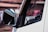 A close-up view of the side mirror of a white vehicle, focusing on the reflective surface and part of the window. The background is blurred, with hints of pink and brown hues, suggesting an outdoor setting.