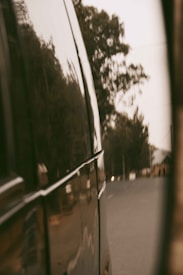 A reflective surface of a vehicle shows a blurred view of a road surrounded by trees. The angle suggests looking out from the side of a van or bus, giving a sense of travel or movement.