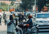 A photographer with a large camera lens is capturing a scene on a busy street. Nearby, a person on a motorcycle, wearing a bright red helmet, passes by. Several other individuals and vehicles are seen in the background, suggesting an urban setting with active movement.