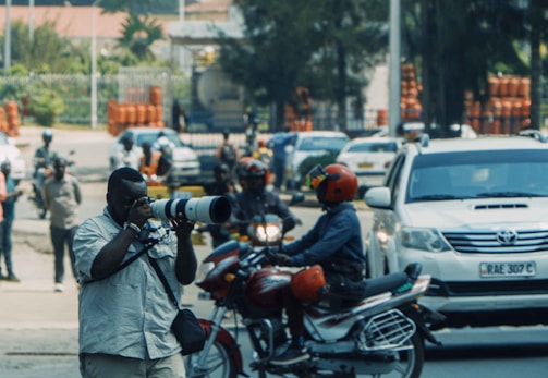 A photographer with a large camera lens is capturing a scene on a busy street. Nearby, a person on a motorcycle, wearing a bright red helmet, passes by. Several other individuals and vehicles are seen in the background, suggesting an urban setting with active movement.