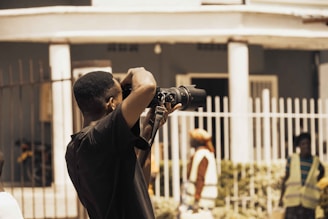 A person is taking a photograph with a professional camera. The scene is outdoors in front of a building with white columns and a fence. In the background, a few people wearing reflective vests are visible, suggesting a street or construction setting. The lighting is natural, with warm tones.