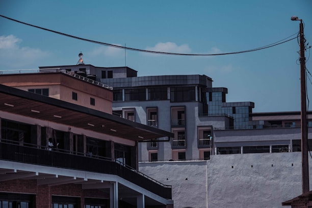 An urban scene featuring modern buildings with geometric architectural design. The structures are primarily made of concrete and glass, with visible balconies and windows. Overhead wires and a utility pole are present, adding to the urban feel. The sky is clear with a few clouds, suggesting it is daytime.