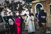 A group of individuals, including clergy members such as a person in a pink ceremonial robe and a nun, standing next to a brick building. Several vehicles and trees are visible in the background. One person is holding a camera and wearing a vest with 'Fr. Ramon KAJUGA TVET School' written on it.