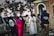 A group of individuals, including clergy members such as a person in a pink ceremonial robe and a nun, standing next to a brick building. Several vehicles and trees are visible in the background. One person is holding a camera and wearing a vest with 'Fr. Ramon KAJUGA TVET School' written on it.