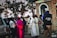 A group of individuals, including clergy members such as a person in a pink ceremonial robe and a nun, standing next to a brick building. Several vehicles and trees are visible in the background. One person is holding a camera and wearing a vest with 'Fr. Ramon KAJUGA TVET School' written on it.