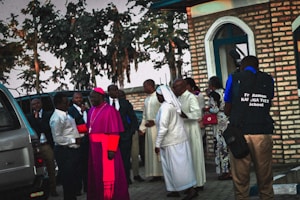 A group of individuals, including clergy members such as a person in a pink ceremonial robe and a nun, standing next to a brick building. Several vehicles and trees are visible in the background. One person is holding a camera and wearing a vest with 'Fr. Ramon KAJUGA TVET School' written on it.