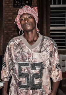 A person stands indoors wearing a red and white checkered headscarf along with a camouflage-patterned sports jersey featuring the number 52 and the word 'Packers'. The background is composed of brick walls and vertical bars.