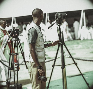 A person in a light vest is standing next to two tripods, likely used for filming or photography. The background is filled with people dressed in white under a tent. The scene seems to be outdoors with a cordoned-off area.