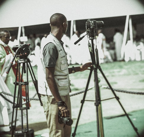 A person in a light vest is standing next to two tripods, likely used for filming or photography. The background is filled with people dressed in white under a tent. The scene seems to be outdoors with a cordoned-off area.