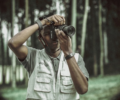 A person is engaged in photography, holding a Canon camera to their eye while wearing a multimedia vest. The background consists of blurred trees, creating an outdoor setting.