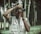 A person is engaged in photography, holding a Canon camera to their eye while wearing a multimedia vest. The background consists of blurred trees, creating an outdoor setting.
