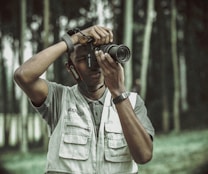 A person is engaged in photography, holding a Canon camera to their eye while wearing a multimedia vest. The background consists of blurred trees, creating an outdoor setting.
