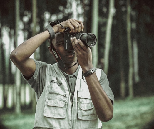 A person is engaged in photography, holding a Canon camera to their eye while wearing a multimedia vest. The background consists of blurred trees, creating an outdoor setting.
