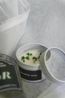 A small tin candle with a white wax surface is adorned with green floral elements. The lid is slightly off, placed on a textured white fabric. There's a blurred glass bottle featuring the letters 'AR'. The date 'septembrie 2023' is visible on the candle's label.