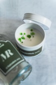 A white candle in a round tin container with a lid that's slightly ajar, featuring small green flowers on its surface. The tin has a label with '17 Septembrie 2023' written on it. Nearby, there is a transparent bottle with a dark label including the letters 'MR'. The background consists of a soft, light textured fabric.