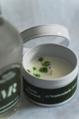 A white candle in a tin container decorated with small green plants around the wick. The container's lid is placed beside it, and there's a blurred bottle in the foreground with text partially visible.