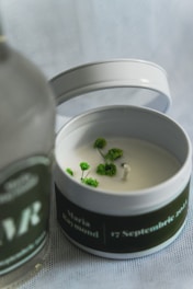 A white candle in a tin container decorated with small green plants around the wick. The container's lid is placed beside it, and there's a blurred bottle in the foreground with text partially visible.