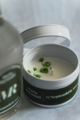 A white candle in a tin container decorated with small green plants around the wick. The container's lid is placed beside it, and there's a blurred bottle in the foreground with text partially visible.