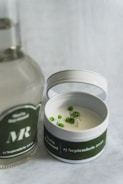 A round candle in a tin container with a green label is positioned next to a clear bottle with a similar green label. The candle has small green decorative elements on its surface, and the labels feature white text including a date and names. The background is a light, textured fabric.