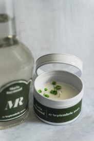 A round candle in a tin container with a green label is positioned next to a clear bottle with a similar green label. The candle has small green decorative elements on its surface, and the labels feature white text including a date and names. The background is a light, textured fabric.