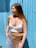 A woman stands against a bright blue corrugated wall, wearing light blue denim jeans and a white cropped top. She has a beige suede fringe bag across her waist and long, straight hair. Her accessories include layered necklaces and small rectangular sunglasses with blue hues.