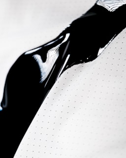 A close-up view of a smooth and shiny black liquid flowing over a white surface with small polka dots. The liquid creates a glossy texture with reflections of light.