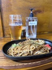A glass of beer and a sizzling plate of yakisoba are on a wooden table. There is a condiment bottle and a pump bottle in the background.