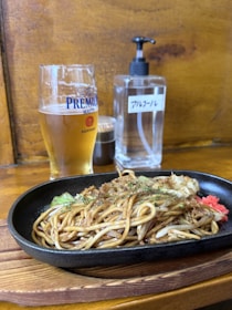 A glass of beer and a sizzling plate of yakisoba are on a wooden table. There is a condiment bottle and a pump bottle in the background.