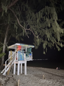 A lifeguard station stands on a sandy beach, illuminated at night under a canopy of tree branches. The structure is elevated on stilts, with a flight of stairs leading up to it. Bright signage covers the front, and there are some lifeguard equipment, including a rescue board and floatation devices. The background reveals a dark sea and distant city lights along the horizon. Two people are sitting on the sand.