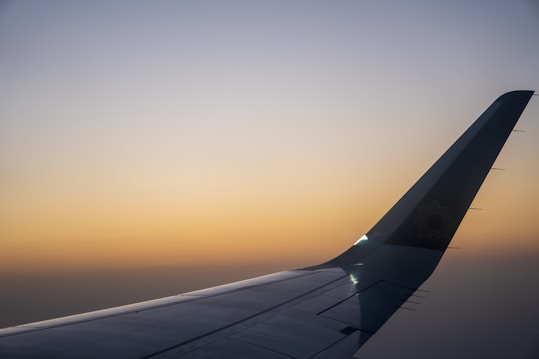A sleek airplane wing extends into the distance against a backdrop of a serene sunset. The sky displays a gradient from a warm orange near the horizon to a cooler blue at the top, creating a tranquil atmosphere.