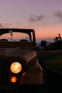 A vintage jeep is parked with its headlights on during an early evening. The sky is filled with hues of orange and purple, indicating sunset. There are some trees visible in the background, contributing to the serene atmosphere.