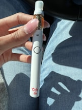 A hand holding a vape pen with a white body and brown liquid cartridge, wearing light blue denim jeans. The pen has a 'Cake' logo near the bottom and is held against a background of car interior.