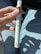 A hand holding a vape pen with a white body and brown liquid cartridge, wearing light blue denim jeans. The pen has a 'Cake' logo near the bottom and is held against a background of car interior.