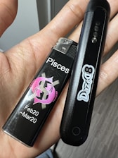 A person's hand is holding a black lighter with a pink Pisces symbol, along with the date range Feb20 - Mar20, and a black electronic device with the brand name 'DazeD' printed on it.
