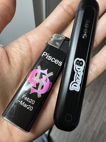 A person's hand is holding a black lighter with a pink Pisces symbol, along with the date range Feb20 - Mar20, and a black electronic device with the brand name 'DazeD' printed on it.