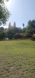 A bright yellow church with twin towers is perched on a hill, surrounded by lush green trees. In the foreground, there is an open grassy area with a few people walking and some signs posted along a pathway.