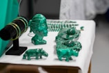 A collection of green crystal or gemstone carvings and jewelry displayed on a table. The centerpiece is a bust of a face with intricate details next to various animal figures and bracelets on a black stand. The objects are placed on a white surface, accentuating their green color.