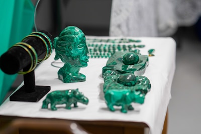 A collection of green crystal or gemstone carvings and jewelry displayed on a table. The centerpiece is a bust of a face with intricate details next to various animal figures and bracelets on a black stand. The objects are placed on a white surface, accentuating their green color.