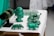 A collection of green crystal or gemstone carvings and jewelry displayed on a table. The centerpiece is a bust of a face with intricate details next to various animal figures and bracelets on a black stand. The objects are placed on a white surface, accentuating their green color.