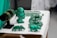 A collection of green crystal or gemstone carvings and jewelry displayed on a table. The centerpiece is a bust of a face with intricate details next to various animal figures and bracelets on a black stand. The objects are placed on a white surface, accentuating their green color.