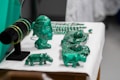 A collection of green crystal or gemstone carvings and jewelry displayed on a table. The centerpiece is a bust of a face with intricate details next to various animal figures and bracelets on a black stand. The objects are placed on a white surface, accentuating their green color.