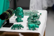 A collection of green crystal or gemstone carvings and jewelry displayed on a table. The centerpiece is a bust of a face with intricate details next to various animal figures and bracelets on a black stand. The objects are placed on a white surface, accentuating their green color.