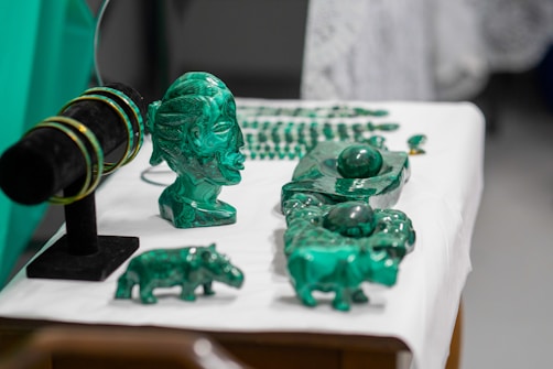 A collection of green crystal or gemstone carvings and jewelry displayed on a table. The centerpiece is a bust of a face with intricate details next to various animal figures and bracelets on a black stand. The objects are placed on a white surface, accentuating their green color.