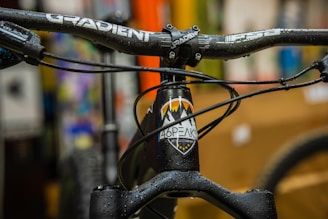 A close-up view of a bicycle's handlebar and front frame, with the brand name 'Gradient' visible on the handlebar. The frame has a logo with mountains and raindrop designs. Water droplets are seen on the handlebars and frame, indicating a wet or rainy environment. The background is blurred with various colors, suggesting an indoor setting or store.
