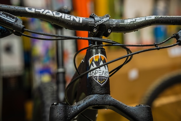 A close-up view of a bicycle's handlebar and front frame, with the brand name 'Gradient' visible on the handlebar. The frame has a logo with mountains and raindrop designs. Water droplets are seen on the handlebars and frame, indicating a wet or rainy environment. The background is blurred with various colors, suggesting an indoor setting or store.