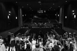 A theater with a full stage, thick curtains on the sides, and dim lighting creating a dramatic atmosphere. The audience consists of people gathered closely, dressed formally, facing the stage. The setting appears elegant with classical architecture.