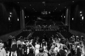 A theater with a full stage, thick curtains on the sides, and dim lighting creating a dramatic atmosphere. The audience consists of people gathered closely, dressed formally, facing the stage. The setting appears elegant with classical architecture.